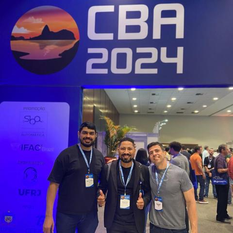 Professores Danilo e Ermeson e o egresso Edson Moura posam para foto em frente À entrada do CBA 2024